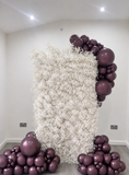 A decorative party backdrop setup featuring a tall rectangular panel covered in dense white faux flowers, framed by clusters of metallic dark purple balloons on the right side and floor around it, photographed in a neutral indoor room.
